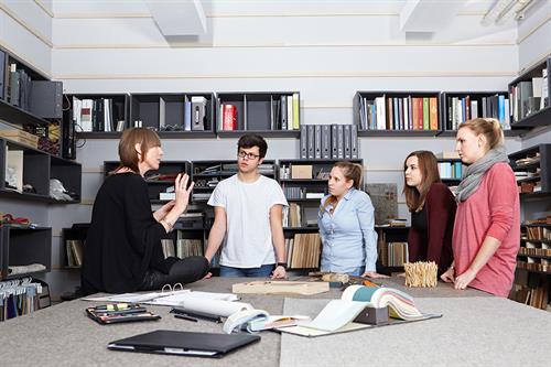 Dieses Bild zeigt eine Professorin und eine Gruppe von Studierenden im Materiallager des Fachbereichs Architektur der Peter Behrens School of Arts.